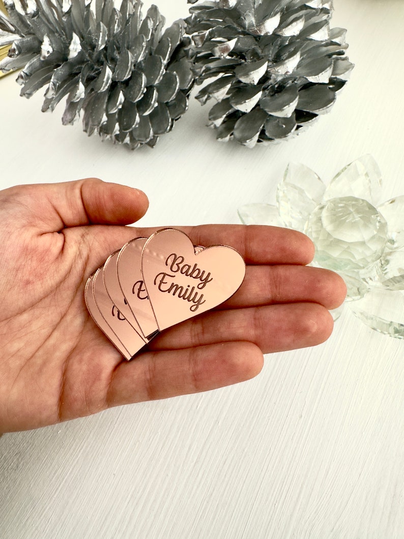 May include: A stack of rose gold heart-shaped acrylic place cards with the text "Baby Emily" engraved on them.