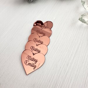 May include: Six rose gold heart-shaped acrylic mirror tiles with the text "Baby" and "Baby Emily" engraved on them.