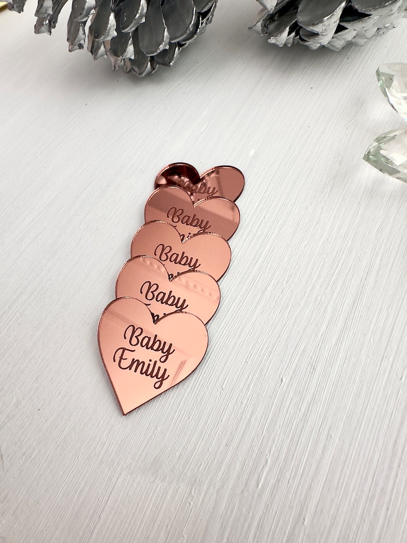 May include: Six rose gold heart-shaped acrylic mirror tiles with the text "Baby" and "Baby Emily" engraved on them.