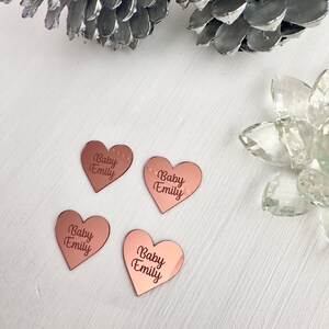 May include: Four rose gold heart-shaped mirror tiles with the text "Baby Emily" engraved on each.