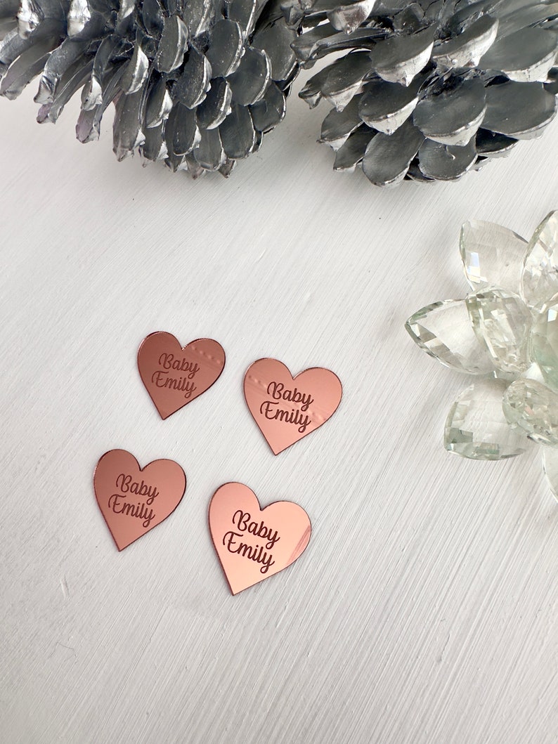 May include: Four rose gold heart-shaped mirror tiles with the text "Baby Emily" engraved on each.