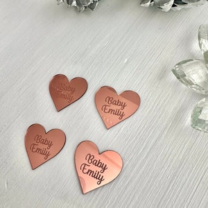 May include: Four rose gold heart-shaped acrylic ornaments with the text "Baby Emily" engraved on each.