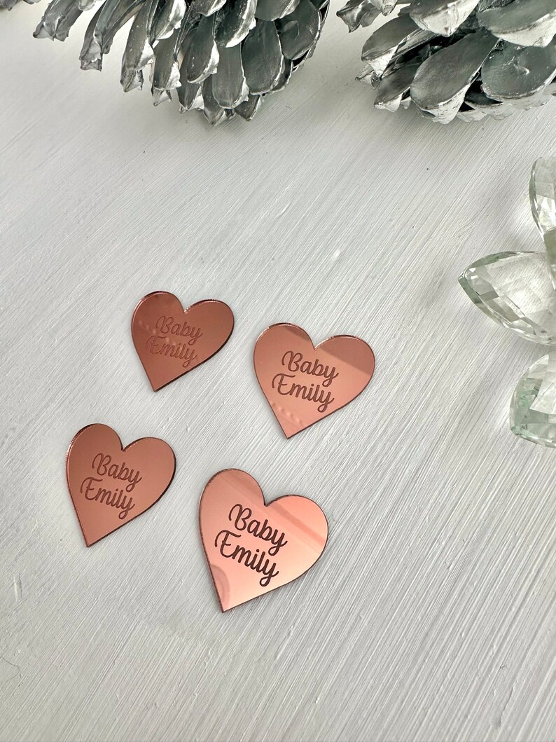May include: Four rose gold heart-shaped acrylic ornaments with the text "Baby Emily" engraved on each.