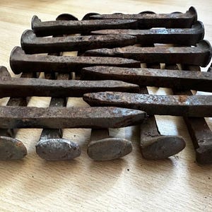 May include: A collection of rusty railroad spikes arranged in a staggered pattern. The spikes are made of metal and have a pointed end. They are all different lengths and have a weathered, aged appearance.