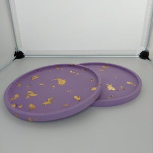 Op de afbeelding: Twee ronde paarse onderzetters met gouden vlokken. De onderzetters zijn gemaakt van hars en hebben een gladde, glanzende afwerking.