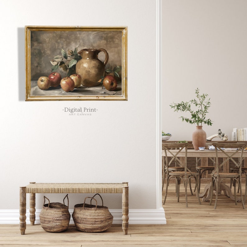 May include: A still life painting of a brown ceramic pitcher and red apples on a table with a white tablecloth. The painting is framed in a gold frame and is hanging on a white wall.