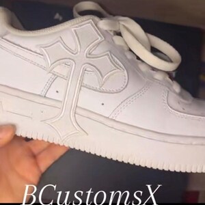 May include: White trainer with a cross design on the side. The shoe has white laces and a thick white sole. The text "BCustomsX" is visible at the bottom of the image.