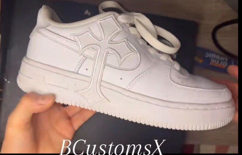May include: White trainer with a cross design on the side. The shoe has white laces and a thick white sole. The text "BCustomsX" is visible at the bottom of the image.