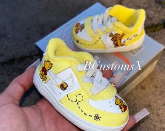Könnte beinhalten: Gelb-weiße Babyschuhe mit Illustrationen von Winnie Puuh und einem Honigtopf. Die Schuhe haben weiße Schnürsenkel und einen gelben gepolsterten Kragen. Das Design beinhaltet eine Biene und gepunktete Linien. Der Text "BCustoms X" ist sichtbar.
