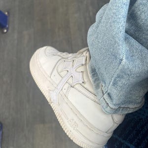 May include: White leather trainer with a cross design overlay. The shoe features the word "AIR" on the side and is paired with light blue denim jeans. The trainer has a thick white sole.