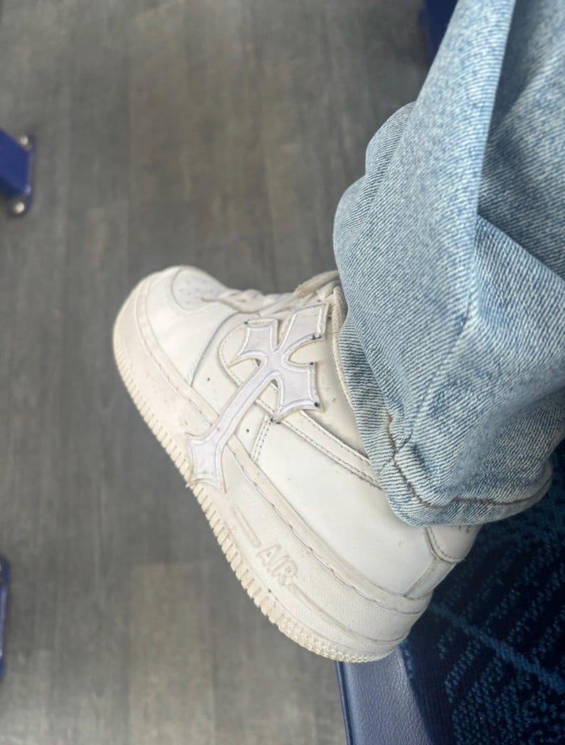 May include: White leather trainer with a cross design overlay. The shoe features the word "AIR" on the side and is paired with light blue denim jeans. The trainer has a thick white sole.