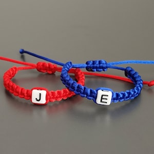 May include: Two friendship bracelets, one red and one blue, with white beads that spell out the letters "J" and "E".