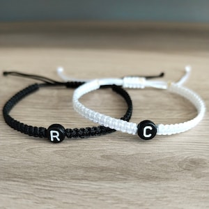 May include: Two friendship bracelets with black and white cord, one with a black bead with the letter "R" and the other with a black bead with the letter "C".