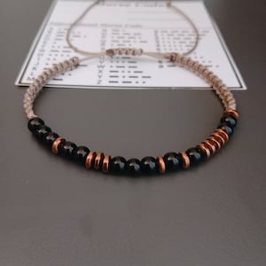 May include: A black and copper beaded bracelet with a beige cord. The bracelet is made with round black beads and copper spacer beads.