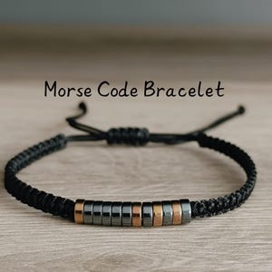May include: A black cord bracelet with a row of black and gold beads. The bracelet is adjustable and has a sliding knot. The bracelet is on a wooden surface. Morse Code Bracelet