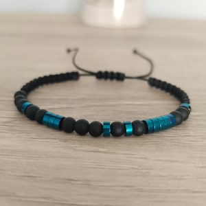 May include: A beaded bracelet featuring matte black spherical beads interspersed with metallic teal cylindrical spacers. The bracelet is constructed with a black cord and includes an adjustable sliding knot closure mechanism at the back.