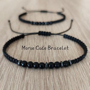 May include: Two black Morse code bracelets are displayed on a wooden surface. The bracelets feature black beads and a braided cord. The text "Morse Code Bracelet" is visible in the image.