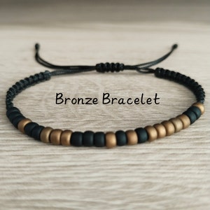 May include: A bracelet featuring alternating matte black and metallic bronze beads on a black cord with an adjustable sliding knot closure. The text Bronze Bracelet is centered above the jewelry piece, which is constructed from small round beads.
