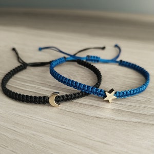 May include: Two friendship bracelets, one black and one blue, with a gold crescent moon charm on the black bracelet and a gold star charm on the blue bracelet.