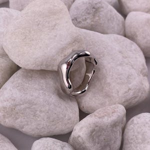 Könnte beinhalten: Ein silberfarbener Ring mit organischem, fließendem Design. Der Ring hat eine glatte, polierte Oberfläche und eine unregelmäßige Form. Der Ring wird auf einem Bett aus weißen, glatten Steinen präsentiert, was einen Kontrast in Textur und Farbe erzeugt.