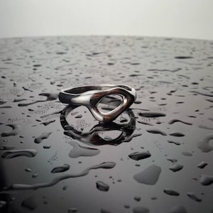 Op de afbeelding: Een zilveren ring met een open, abstract ontwerp. De ring is geplaatst op een donker, reflecterend oppervlak bedekt met waterdruppels, waardoor een contrast ontstaat tussen het glanzende metaal en de natte achtergrond. De vorm van de ring is organisch en modern.