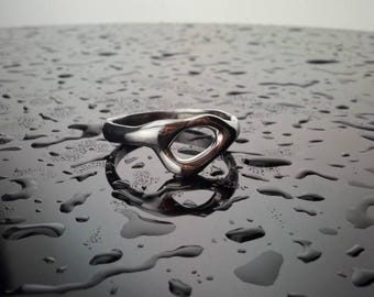 May include: A silver ring with an open, abstract design. The ring is set against a dark, reflective surface covered in water droplets, creating a contrast between the shiny metal and the wet background. The ring's shape is organic and modern.
