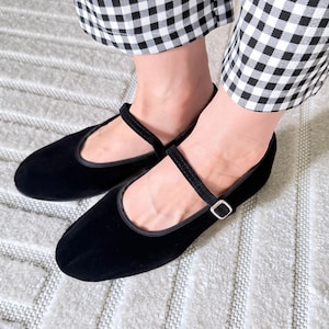 May include: Black velvet Mary Jane flats with a strap and buckle closure. The shoes have a rounded toe and a low heel. The shoes are shown with black and white checkered pants.