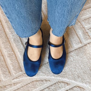 May include: A pair of navy blue Mary Jane shoes with a strap and buckle, viewed from above. The shoes have a shiny finish and black trim. They are worn with blue jeans. The background is a beige rug with a geometric pattern.