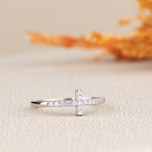 Pu&ograve; includere: Un anello d'argento con un piccolo design a croce incastonato con pietre di zircone cubico trasparenti.