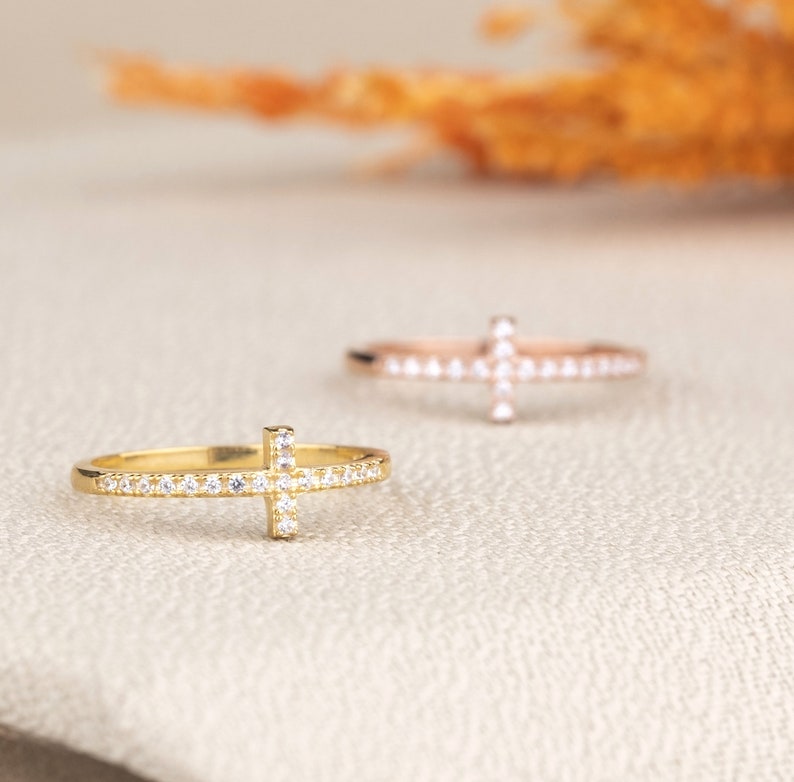 Pu&ograve; includere: Due anelli d'oro con un design a croce incastonati con piccole pietre trasparenti. Gli anelli sono su una superficie testurizzata di colore marrone chiaro.