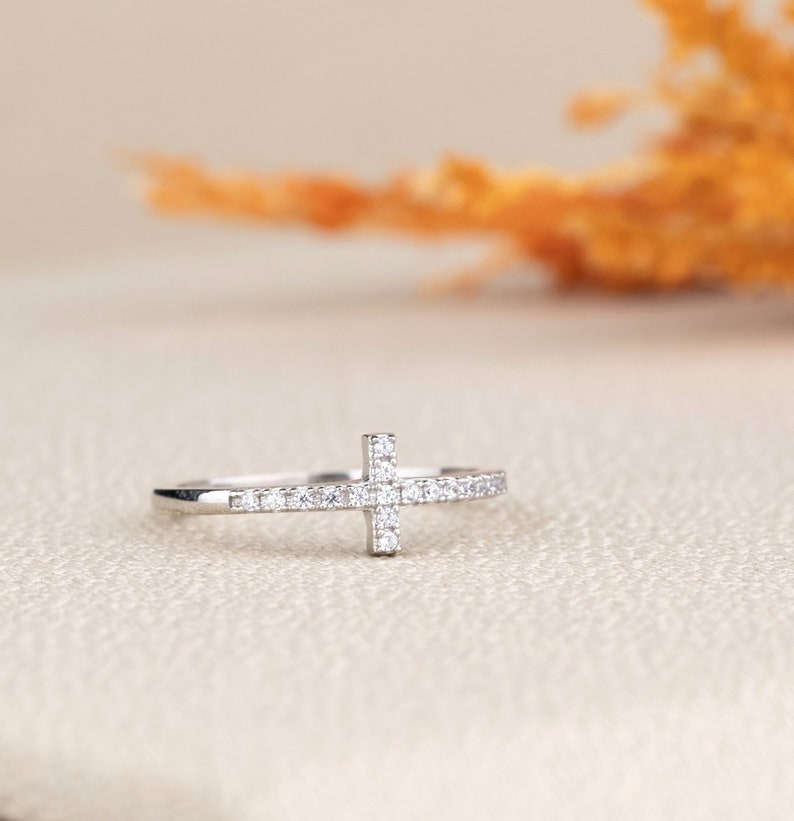 Pu&ograve; includere: Un anello d'argento con un piccolo design a croce incastonato con pietre di zircone cubico trasparenti.