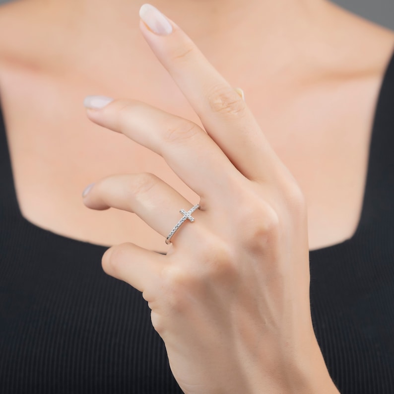 Pu&ograve; includere: Un anello d'argento con un piccolo design a croce incastonato con pietre di zircone cubico trasparenti.