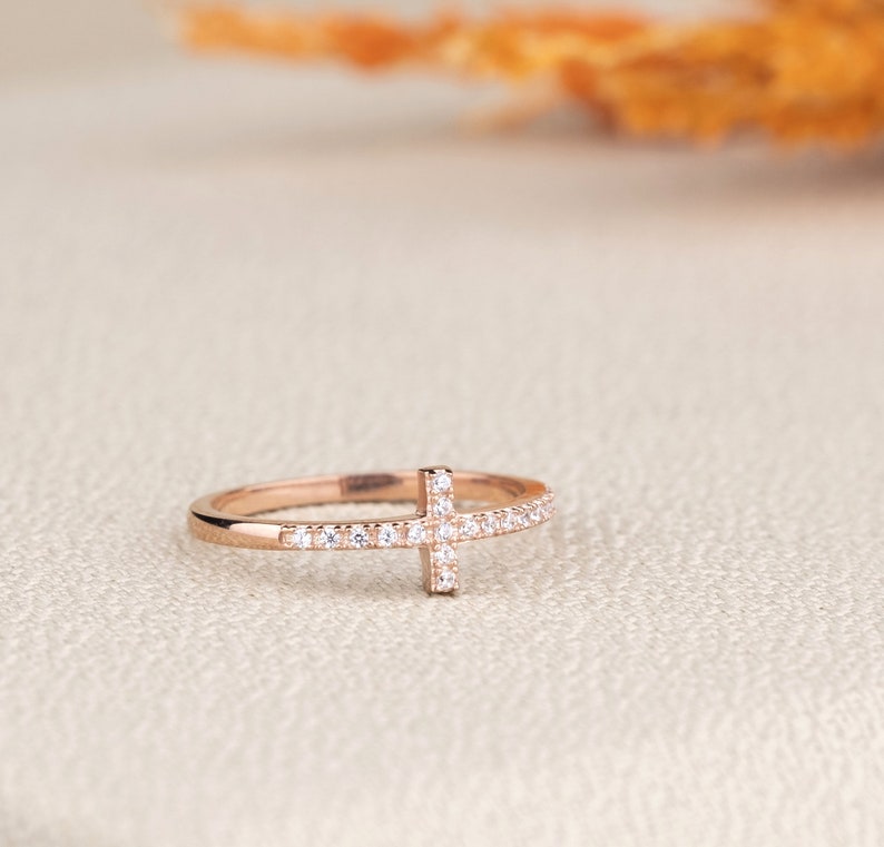 Pu&ograve; includere: Un anello in oro rosa con un piccolo design a croce incastonato con pietre di zircone cubico trasparenti.