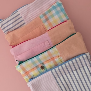 May include: Several zippered pouches in a variety of patterns and colours. The pouches combine striped, checked, and solid fabric panels in pastel shades such as pink, peach, and blue. Each pouch has a contrasting colour zip.