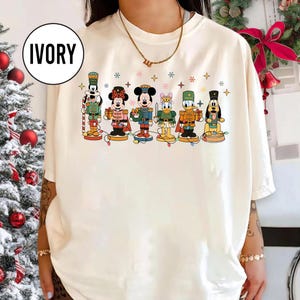 May include: Ivory-colored t-shirt featuring a colorful graphic of Disney characters dressed as nutcrackers. The characters include Mickey Mouse, Minnie Mouse, Donald Duck, and Pluto, with snowflakes and stars. The shirt has a round neckline.