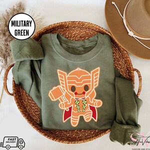 May include: A green sweatshirt with a cartoon gingerbread Thor design. The design features a gingerbread man with a red cape and a hammer. The text "Military Green" is displayed in a white box.