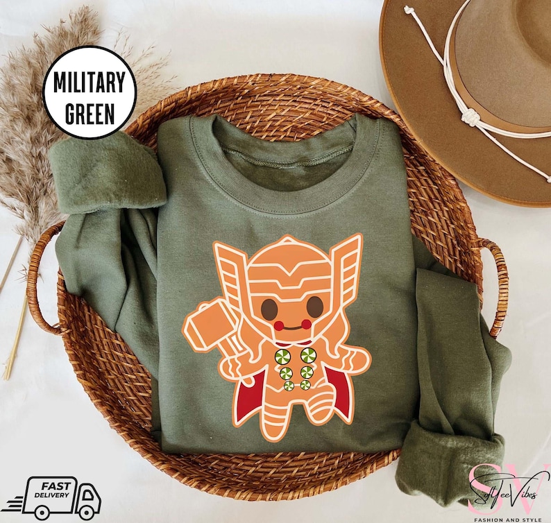 May include: A green sweatshirt with a cartoon gingerbread Thor design. The design features a gingerbread man with a red cape and a hammer. The text "Military Green" is displayed in a white box.