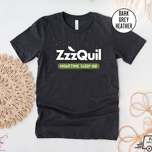 May include: Dark grey heather t-shirt with the text "ZzzQuil NIGHTTIME SLEEP-AID" in white and green. The shirt has a crew neck and short sleeves. A white circle with the words "DARK GREY HEATHER" is in the upper right corner.