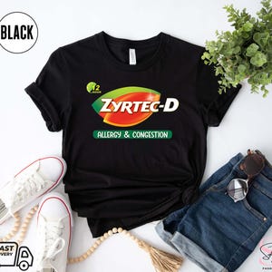 May include: Black t-shirt with the text "ZYRTEC-D ALLERGY & CONGESTION" in green, red, and white. The shirt is styled with white sneakers, blue denim shorts, and sunglasses. A small plant and a beaded necklace are also in the image.