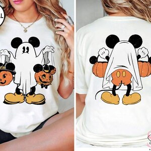 Puede incluir: Camiseta blanca con una ilustraci&oacute;n de estilo vintage de Mickey Mouse vestido de fantasma sosteniendo dos calabazas de jack-o'-lantern. La parte posterior de la camiseta presenta a Mickey Mouse con una s&aacute;bana de fantasma drapeada sobre su espalda.