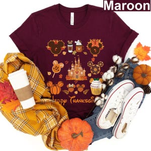 Puede incluir: Una camiseta de color granate con un diseño de temática de Acción de Gracias con Mickey Mouse y sus amigos en Disney World. El diseño incluye a Mickey Mouse, Minnie Mouse, Donald Duck, Goofy y Pluto. El texto "Happy Thanksgiving" está impreso en la camiseta.