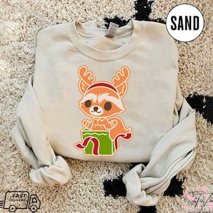 May include: A light beige sweatshirt with a cartoon image of a red fox wearing a Santa hat and holding a green gift box. The fox has a white muzzle and brown eyes. The text "SAND" is in the upper right corner of the image.