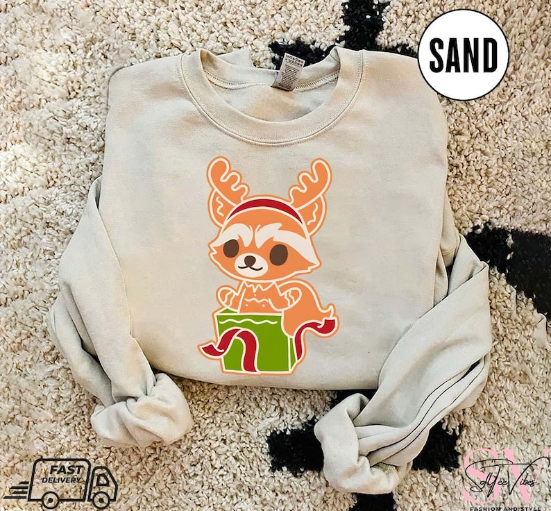 May include: A light beige sweatshirt with a cartoon image of a red fox wearing a Santa hat and holding a green gift box. The fox has a white muzzle and brown eyes. The text "SAND" is in the upper right corner of the image.