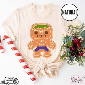 May include: A beige t-shirt with a gingerbread man design. The gingerbread man is green, orange, and purple and resembles the Hulk. The text "NATURAL" is on a white tag.