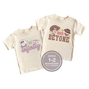 Puede incluir: Dos camisetas color crema con gráficos de dibujos animados. Una presenta a Buzz Lightyear con el texto "To Infinity" en morado. La otra tiene a Woody con el texto "And Beyone" en marrón. Ambas camisetas tienen mangas cortas.