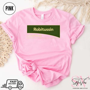 May include: Pink t-shirt with the word "Robitussin" in white text on a dark green rectangle. The text "COUGH RELIEF" is below. The shirt has rolled-up sleeves and a knot at the bottom. A "Pink" circle is in the upper left.