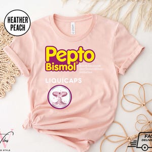 May include: A light pink t-shirt with the Pepto Bismol logo in yellow and purple. The shirt also features the text "LIQUICAPS" and a graphic of a pink capsule. The shirt is from the brand "Softtee Vibes".