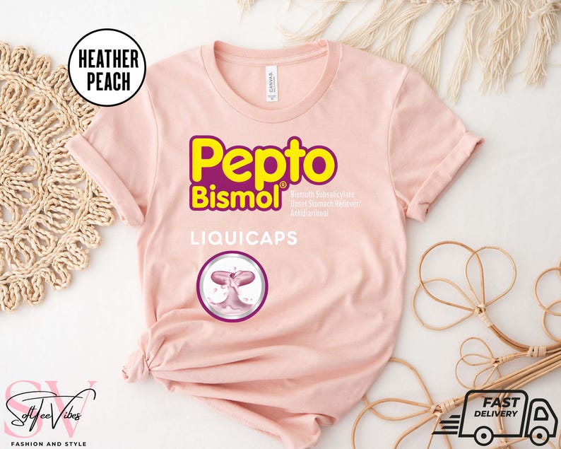 May include: A light pink t-shirt with the Pepto Bismol logo in yellow and purple. The shirt also features the text "LIQUICAPS" and a graphic of a pink capsule. The shirt is from the brand "Softtee Vibes".