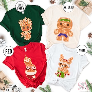 May include: Four different t-shirts with Christmas-themed designs featuring characters from popular movies and TV shows. The shirts are in red, green, white, and natural colors. The designs include a gingerbread man version of the Hulk, a gingerbread man version of Rocket Raccoon, a gingerbread man version of Spider-Man, and a gingerbread man version of Groot.