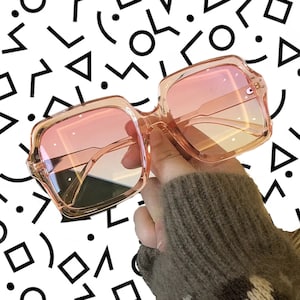 May include: Pink square-shaped sunglasses with a gradient lens. The sunglasses have a thick frame and are made of plastic.